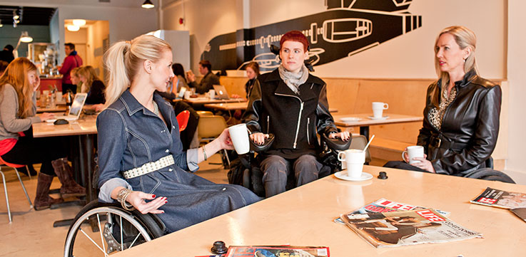 3 women in wheel chairs sitting around a table conversing wearing IZ Adaptive designs